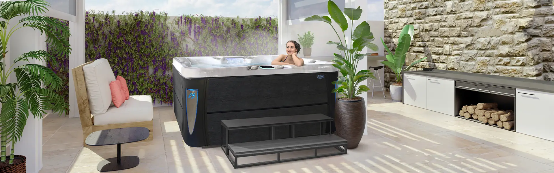 A person lounging in a Cal Spas Excape X hot tub with lime oak cabinet, blue lighted corners and 60-inch steps, in an outdoor living space.